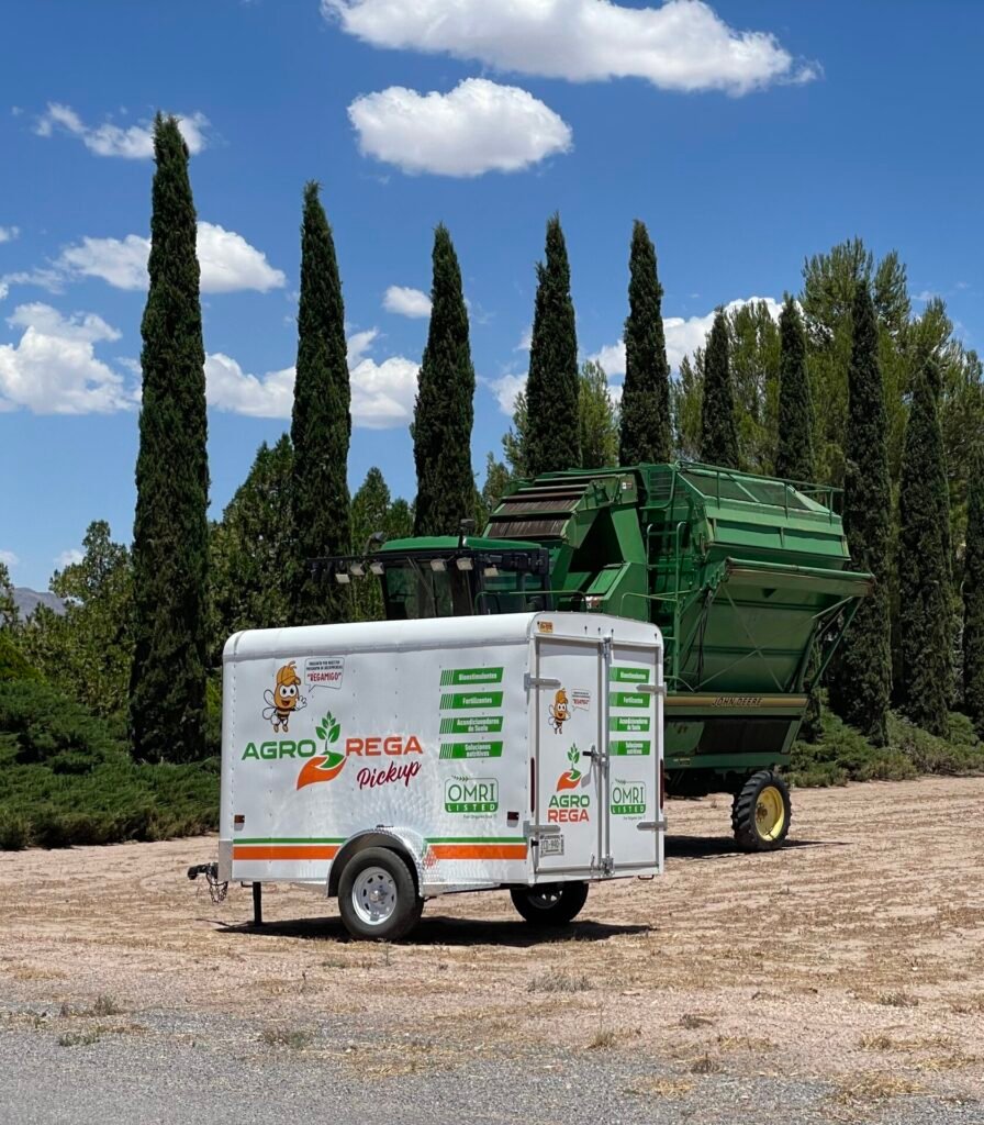 carreta con los logos de Agro Rega "Pickup" enfrente de unos pinos y un tractor John Deere